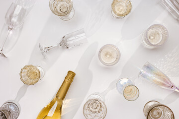 Top view white sparkling wine bottle, set colored glasses wine with sunshine shadow and flare on white background. White wine aesthetic flat lay, trend pattern. Summer still life light alcohol
