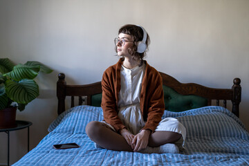 Relaxed teenage girl wearing headphones sitting on bed in lotus pose and looking aside, listen audio content, learning language with podcasts. Millennial woman listening music in leisure time - Powered by Adobe
