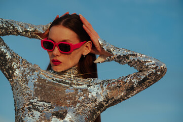 Fashionable confident woman wearing trendy fuchsia color rectangular sunglasses, sequin  turtleneck top, posing outdoor, against blue sky. Close up fashion portrait. Copy, empty space for text - 614468714
