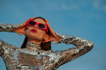 Fashionable confident woman wearing trendy fuchsia color rectangular sunglasses, sequin  turtleneck top, posing outdoor, against blue sky. Close up fashion portrait. Copy, empty space for text - 614468716