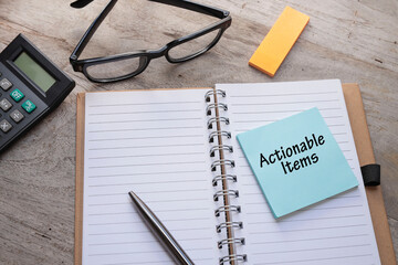 The word Actionable Items is written on the Post-it note in the notebook. A pen, and eyeglasses are on the wooden table 
