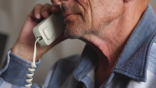 the face of an unshaven elderly man 70-79 years old listens to the interlocutor while talking on the phone. the pensioner holds the handset of a wired telephone. the emotions of the old man