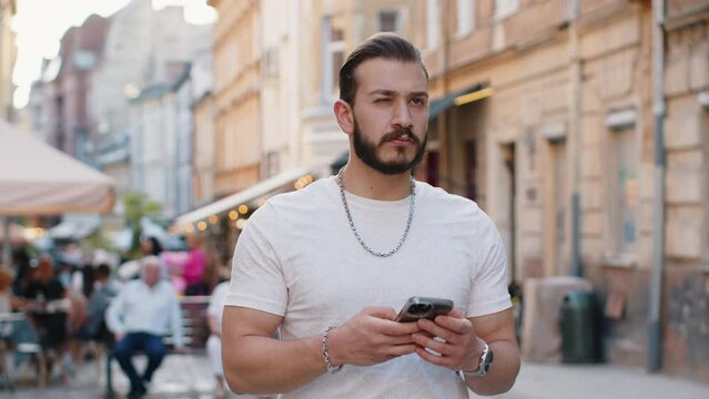 Tourist traveler bearded young man using smartphone typing text messages looking for search a way on map in mobile navigator app outdoors. Guy walking passes by urban sunshine city street background