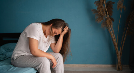 Tired young woman sitting on bed at home having headache, exhausted girl waking up in bedroom,...