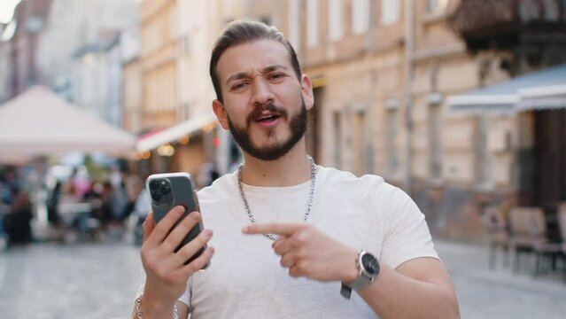 Bearded young man use mobile smartphone celebrating win good message news, lottery jackpot victory, giveaway online outdoors. Happy guy tourist walking in urban sunshine city street. Town lifestyles