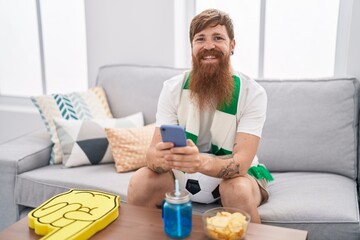 Young redhead man supporting soccer team using smartphone at home