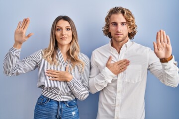 Young couple standing over blue background swearing with hand on chest and open palm, making a loyalty promise oath