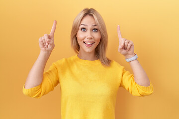 Young caucasian woman wearing yellow sweater smiling amazed and surprised and pointing up with fingers and raised arms.