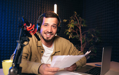 Smiling radio host with headphones reading news from paper into studio microphone at radio station...
