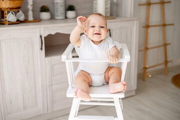 a happy baby in a white cotton bodysuit on a high chair in a bright kitchen wants to eat, a small smiling baby boy or girl