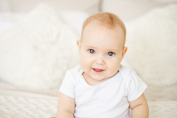 portrait close-up of a baby boy or girl of six months sitting at home on a bed in a bright bedroom and smiling or laughing, a happy newborn in a white bodysuit with blue eyes