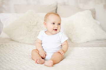 a baby boy or girl of six months is sitting at home on a bed in a bright bedroom and smiling or laughing, a happy newborn in a white bodysuit