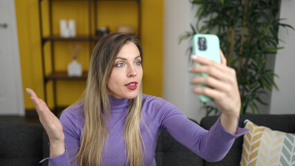 Young blonde woman having video call sitting on sofa at home