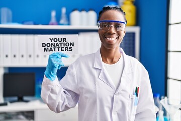 Beautiful black woman working at scientist laboratory holding your donation matters banner looking...