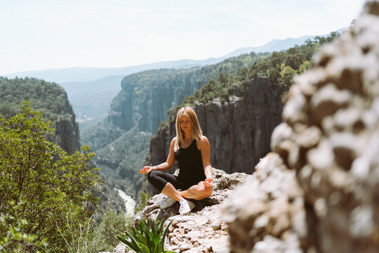 Young beautiful girl woman in black sporty slim outfit meditating on edge rock mountain cliff peak with view on valley canyon. Picture of fit female hiker reached the mountain top.
