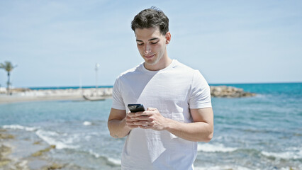 Young hispanic man smiling confident using smartphone at beach