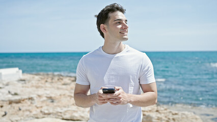 Young hispanic man smiling confident using smartphone at beach