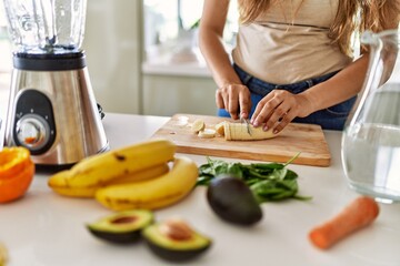 Young beautiful hispanic woman preparing vegetable smoothie with blender cutting banana at the kitchen