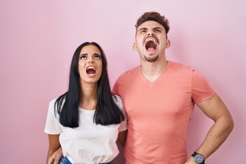 Young hispanic couple standing over pink background angry and mad screaming frustrated and furious, shouting with anger. rage and aggressive concept.