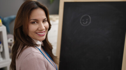 Young beautiful hispanic woman preschool teacher drawing on blackboard at kindergarten
