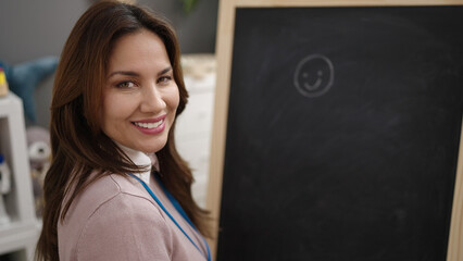 Young beautiful hispanic woman preschool teacher drawing on blackboard at kindergarten