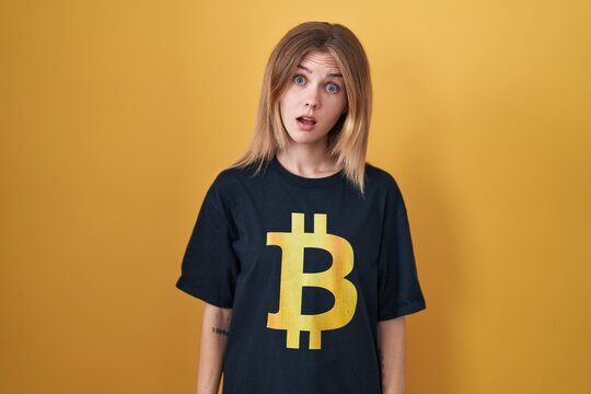 Blonde caucasian woman wearing bitcoin t shirt in shock face, looking skeptical and sarcastic, surprised with open mouth