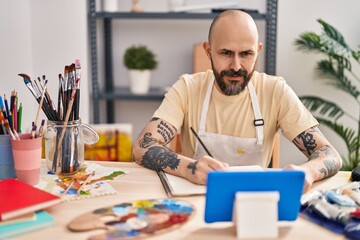Young bald man artist drawing on notebook looking touchpad at art studio