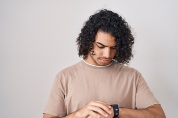 Hispanic man with curly hair standing over white background checking the time on wrist watch, relaxed and confident