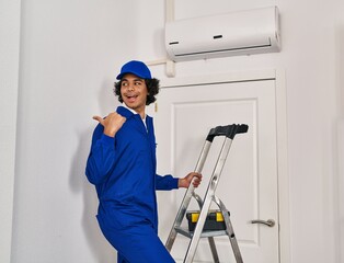Hispanic man with curly hair working at home renovation pointing thumb up to the side smiling happy with open mouth