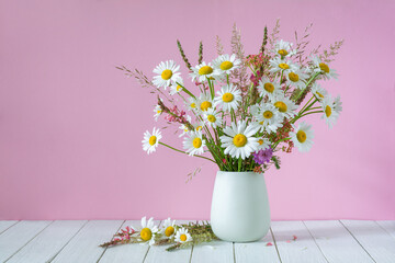 Bouquet of white daisies and other wildflowers in a white vase on a pink background