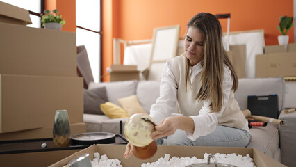 Young beautiful hispanic woman smiling confident unpacking cardboard box at new home