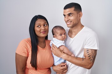 Young hispanic couple with baby standing together over isolated background smiling looking to the side and staring away thinking.