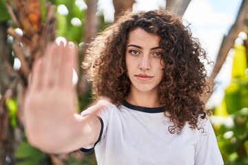 Naklejka premium Hispanic woman with curly hair standing outdoors with open hand doing stop sign with serious and confident expression, defense gesture