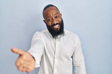 African american man standing over blue background smiling cheerful offering palm hand giving assistance and acceptance.