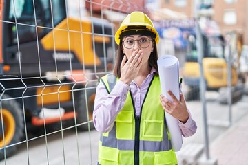 Hispanic girl wearing architect hardhat at construction site covering mouth with hand, shocked and...