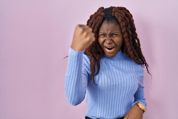 African woman standing over pink background angry and mad raising fist frustrated and furious while...