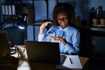 African woman working at the office at night gesturing with hands showing big and large size sign,...