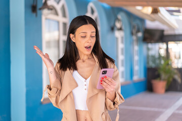 Young woman at outdoors looking at the camera while using the mobile with surprised expression