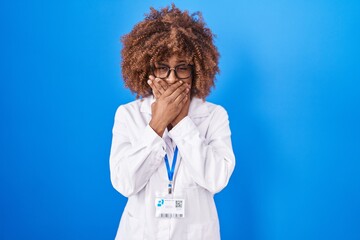 Young hispanic woman with curly hair wearing white coat and id card shocked covering mouth with...