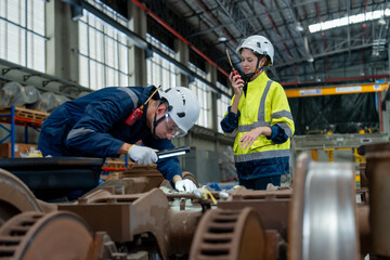 Fototapeta na wymiar Female technician and engineer holding walkie talkie wearing helmet uniform inspects repair and maintenance of electric vehicle wheel and suspension system in electric train maintenance station.