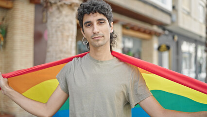 Young hispanic man holding rainbow flag with relaxed expression at street