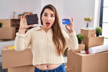 Young hispanic woman moving to a new home holding wallet and credit card afraid and shocked with...