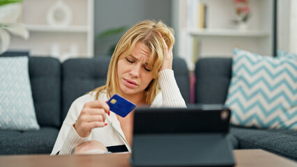 Young blonde woman having video call holding credit card looking upset at home