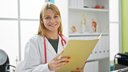 Young blonde woman doctor smiling confident reading medical report at clinic
