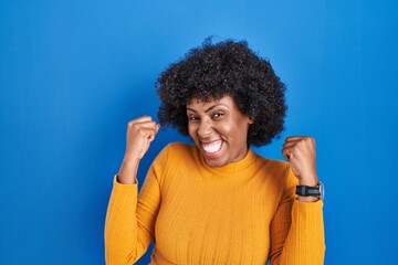 Fototapeta na wymiar Black woman with curly hair standing over blue background celebrating surprised and amazed for success with arms raised and eyes closed. winner concept.
