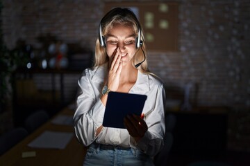 Young blonde woman working at the office at night laughing and embarrassed giggle covering mouth...