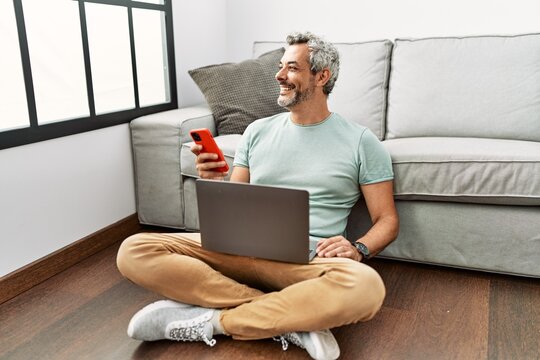 Middle age grey-haired man using smartphone and laptop sitting on floor at home