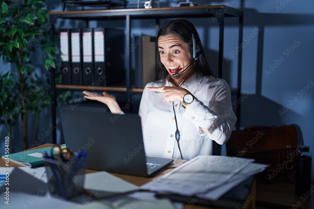 Wall mural Young brunette woman wearing call center agent headset working late at night amazed and smiling to the camera while presenting with hand and pointing with finger.