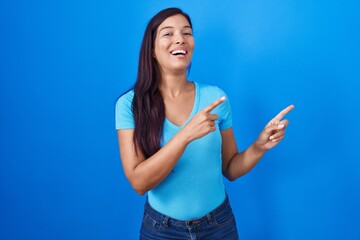 Fototapeta premium Young hispanic woman standing over blue background smiling and looking at the camera pointing with two hands and fingers to the side.