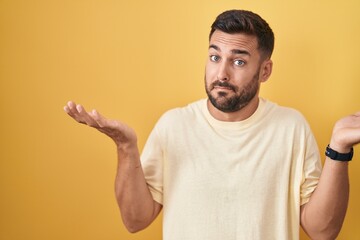 Handsome hispanic man standing over yellow background clueless and confused expression with arms and hands raised. doubt concept.